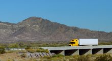 big-rig-long-haul-semi-truck-with-two-flat-bed-semi-trailers-transporting-winding-road-with-bridge-around-mountain-rock